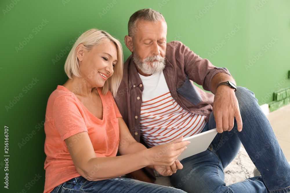 一对成熟的夫妇，带着平板电脑坐在室内彩色墙附近的地板上。搬进新房子