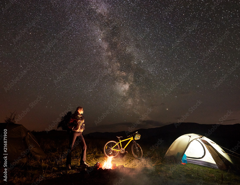 年轻的女自行车手在燃烧的篝火旁的夜间露营中休息，照亮了旅游帐篷，