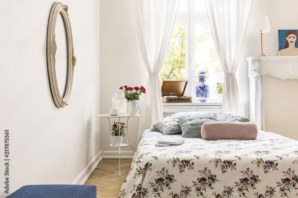 Patterned sheets and pillows on bed in white bedroom interior with mirror and window. Real photo