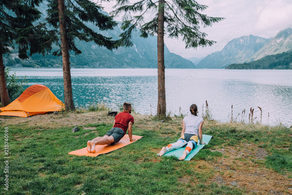 橙色帐篷附近一对运动型情侣的背影，在湖泊和山脉的背景下热身。Th