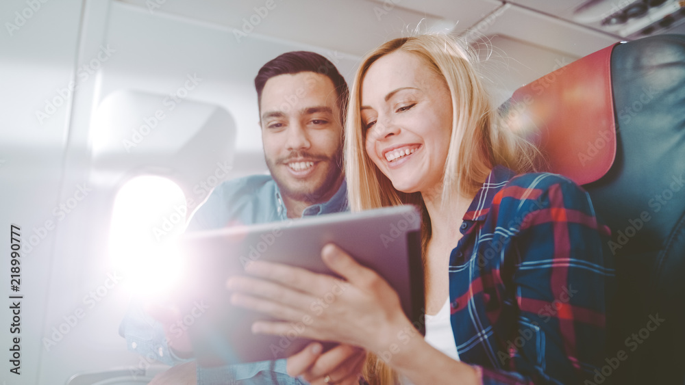 在商用飞机上，美丽的年轻金发女郎和英俊的西班牙裔男性使用平板电脑