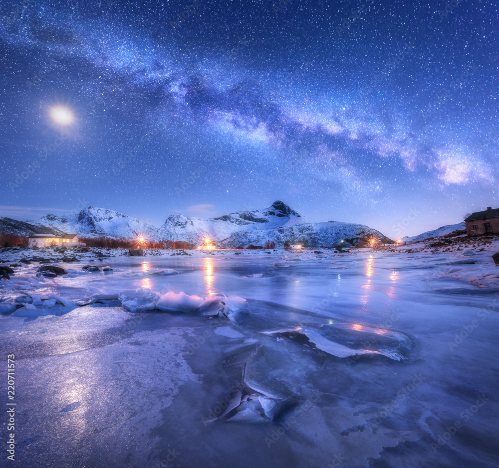Milky Way above frozen sea coast, snow covered mountains and starry sky with moon in winter at night