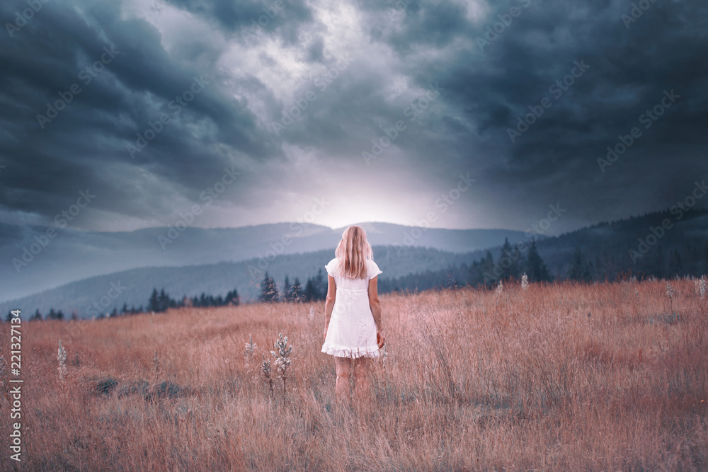 一位身穿白色连衣裙的金发女子站在山顶上，身后是暴风雨般的黑暗天空