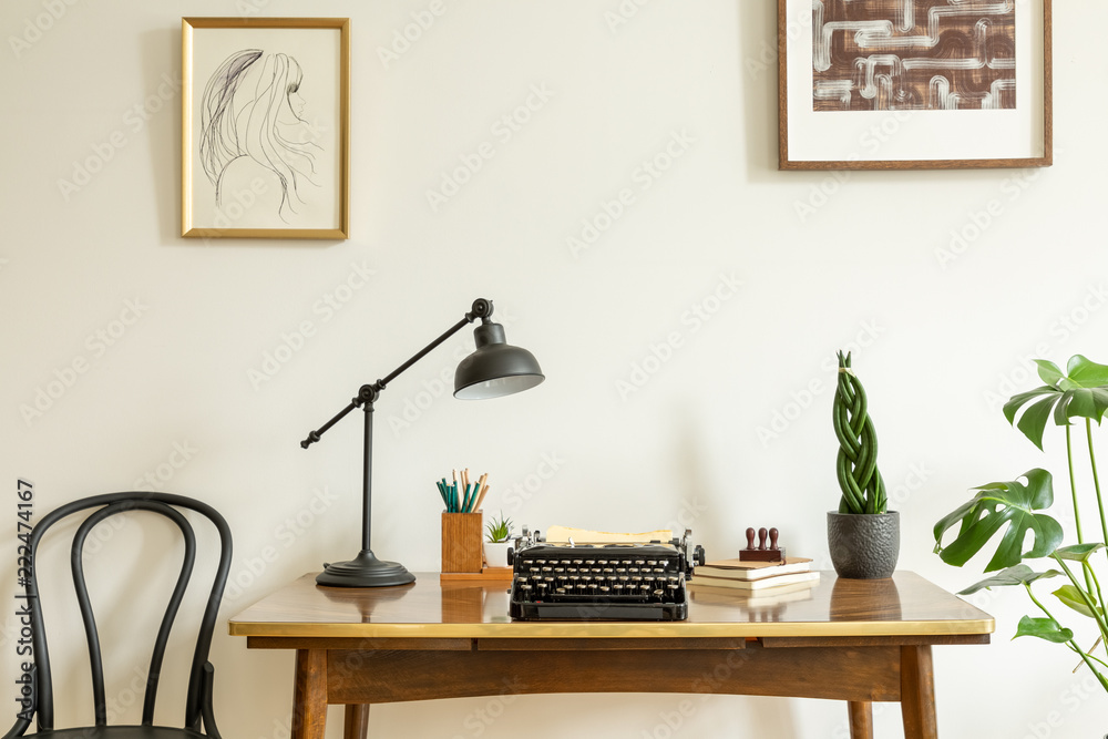 Framed drawing on a white wall above an antique, wooden desk with a vintage, black typewriter in a h