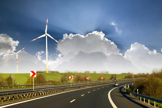 宽阔的高速公路上有移动的汽车延伸到地平线上的绿色山丘，在蓝色的cl上有高高的风力涡轮机