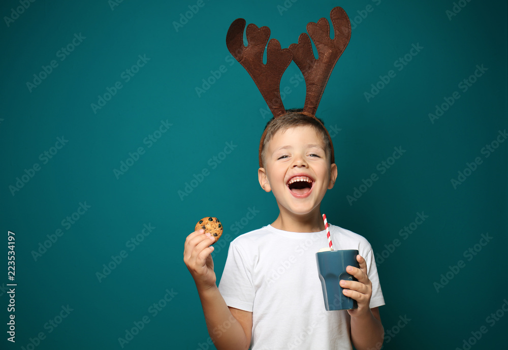 可爱的小男孩，带着玩具驯鹿角，手里拿着一杯彩色背景的热巧克力和饼干