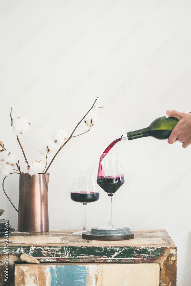 Red wine pouring from bottle into wineglass over rustic kitchen countertop, white background behind,