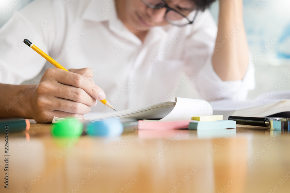 一个十几岁的男子坐在一张木桌旁，在一个博恩特的教室里，在笔记本上看书写字