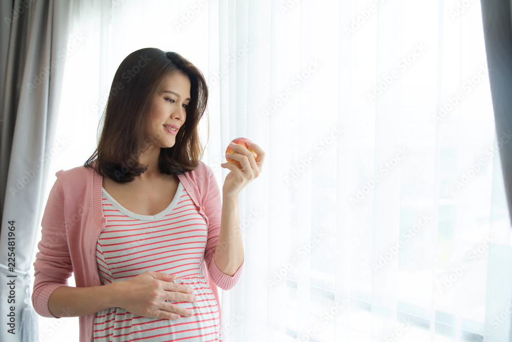 美丽的年轻微笑亚洲粉色连衣裙孕妇在窗前拿着一个苹果。她的右边