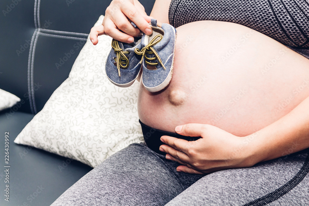 孕妇在家里的沙发上为未出生的婴儿放松并将婴儿鞋放在肚子上