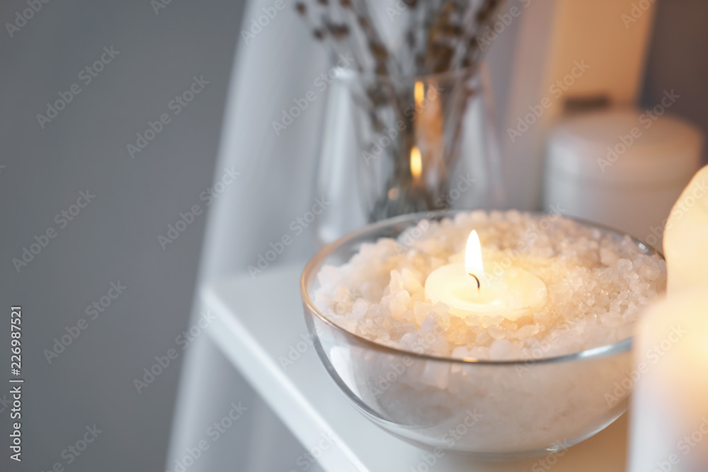Bowl with sea salt and burning candle on shelf indoors