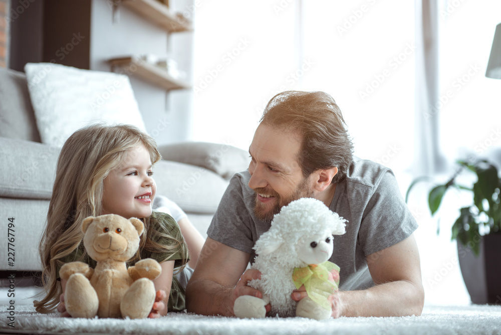 兴奋的女孩在家里玩泰迪熊。她躺在爸爸旁边的地毯上，看起来像一只