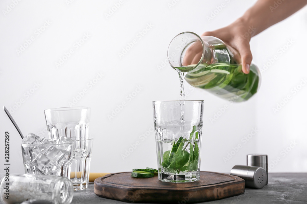一名女子在浅色背景下将美味的新鲜黄瓜水从瓶中倒入玻璃杯