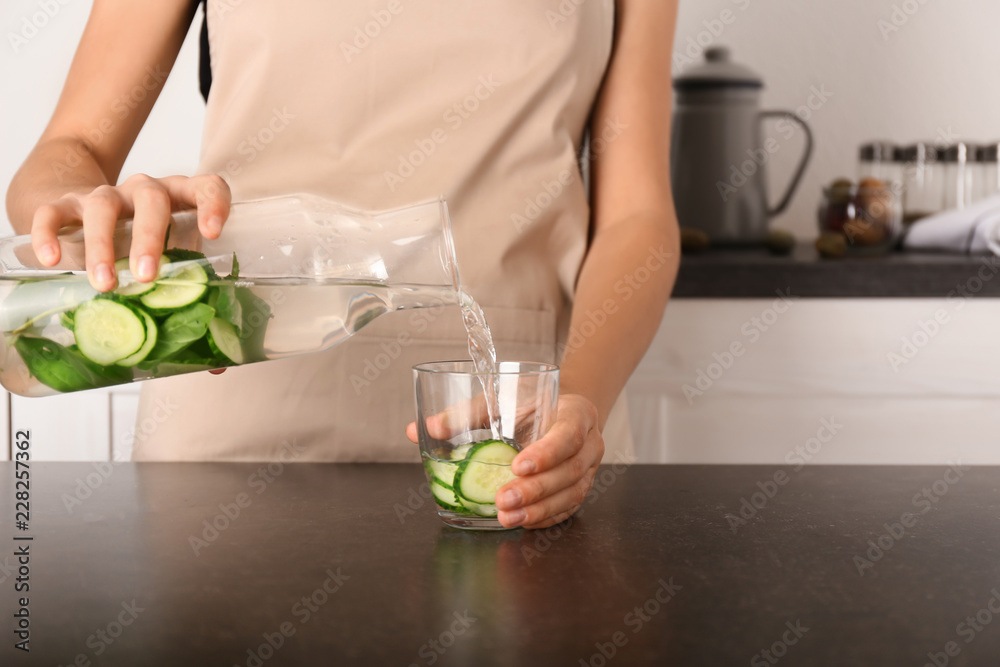 一位女士将美味的新鲜黄瓜水从瓶子倒入黑暗桌子上的玻璃杯