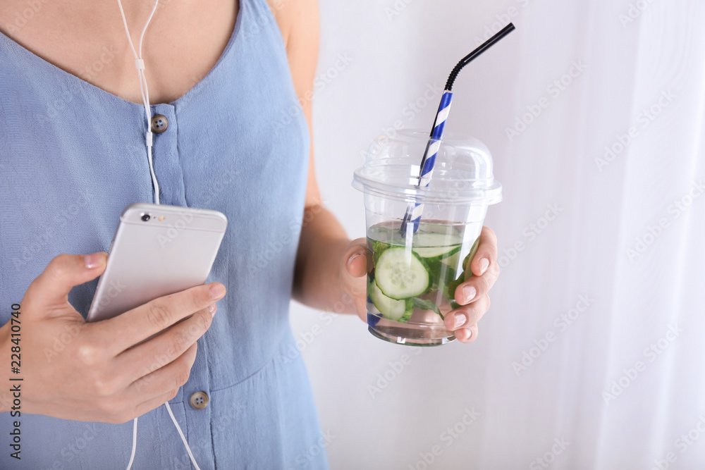 女人拿着装有美味新鲜黄瓜水的塑料杯和手机，特写