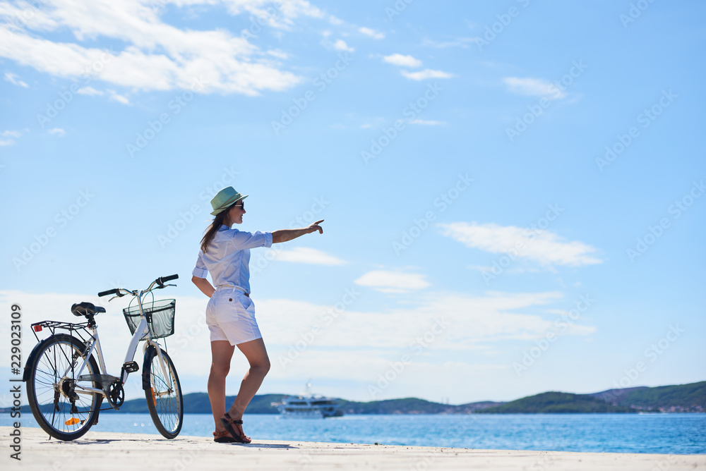 穿着白色紧身衣，戴着遮阳帽的年轻苗条女子站在清澈蓝色的石头人行道上的自行车旁