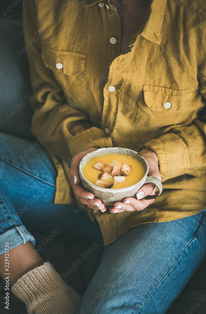 穿着黄色衬衫和牛仔裤的女性坐着，手里拿着一杯秋天温暖的黄色南瓜奶油汤w