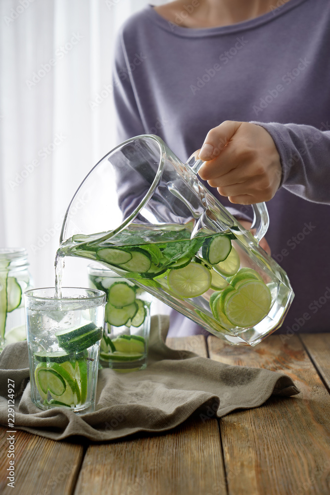 Woman pouring fresh cucumber water into glass from jug