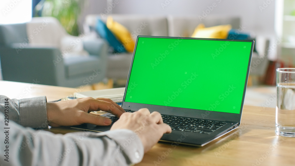 一名男子在舒适的生活中坐在桌子旁使用带有绿色实体屏幕的笔记本电脑的特写
