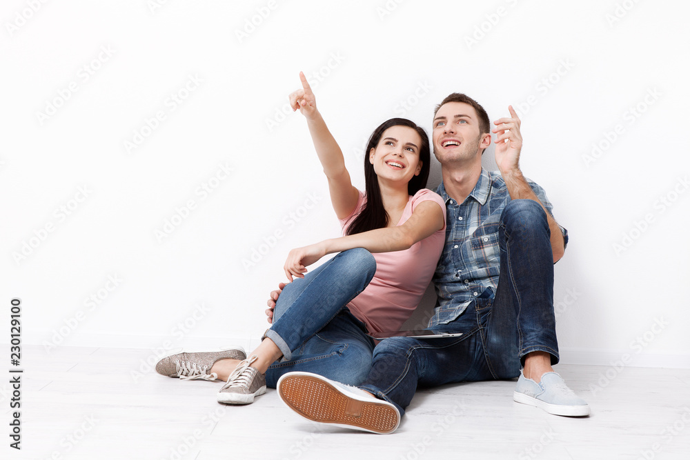 快乐的年轻夫妇坐在地板上，抬头看着他们的新家和家具。实物模型