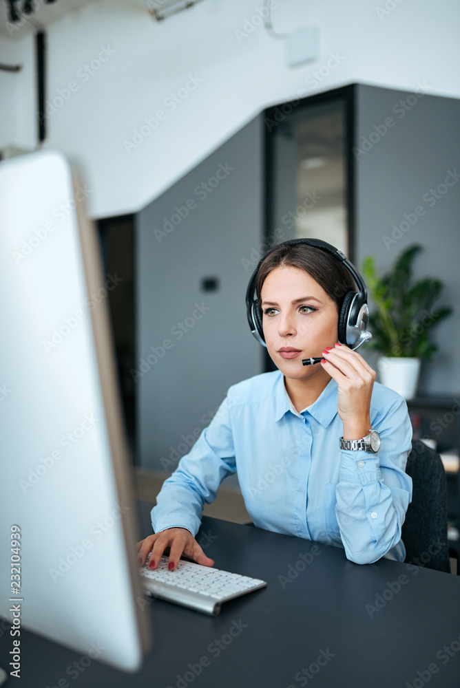 漂亮的年轻女人在现代办公室里戴着耳机和电脑工作。