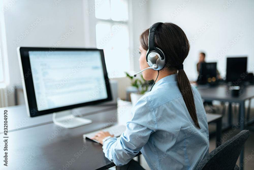 带耳机的女性客户服务代表在电脑上工作。后视图。