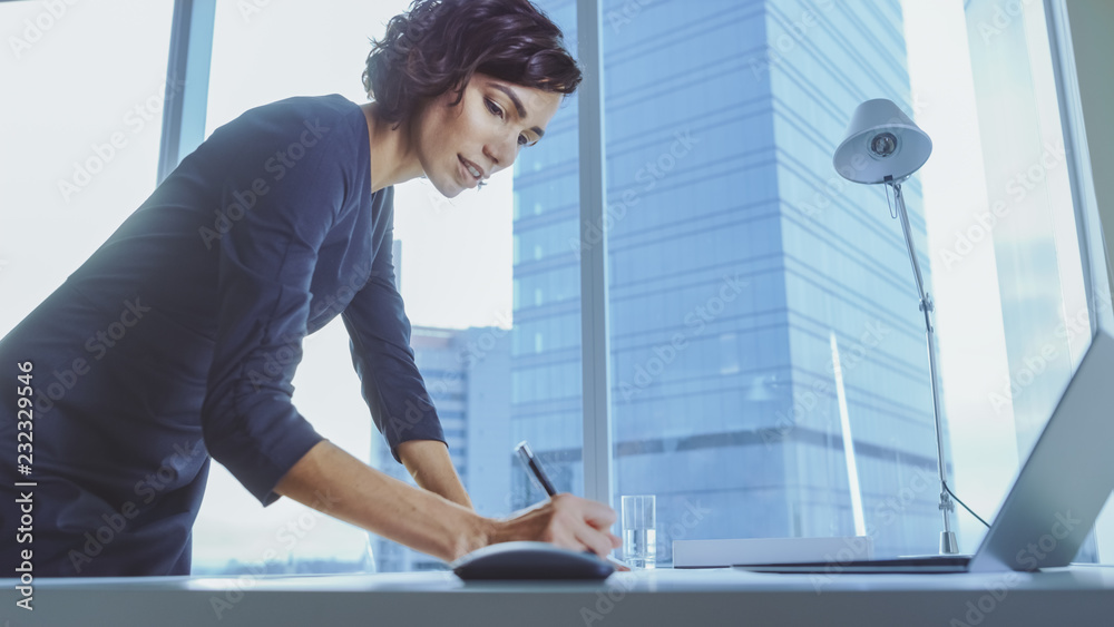自信的女商人靠在办公桌上，在文档中写下信息的低角度镜头