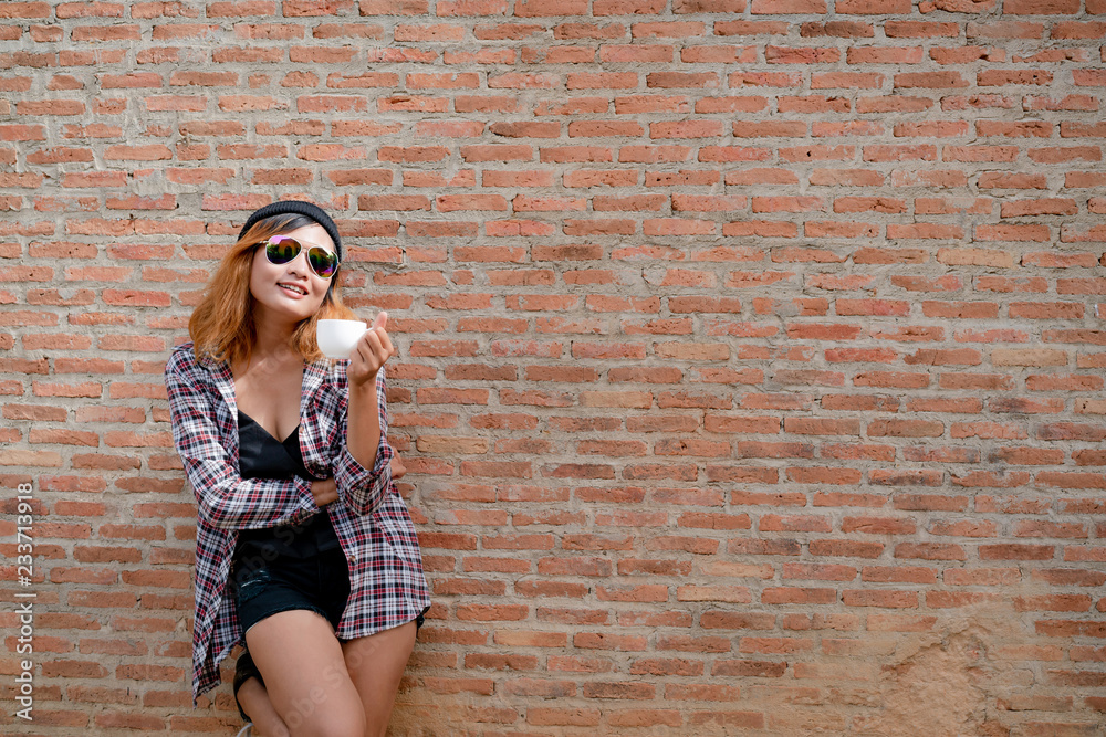 快乐的年轻潮人女人站在镇街的砖墙边，手里拿着咖啡杯。