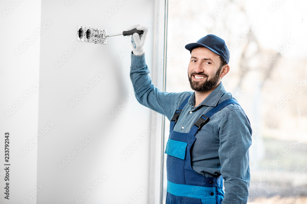 Electrician in uniform mounting electric sockets on the white wall indoors