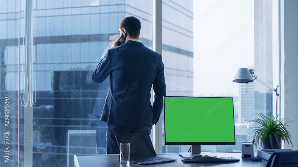 自信的商人在办公室打电话，桌上的个人电脑显示绿色