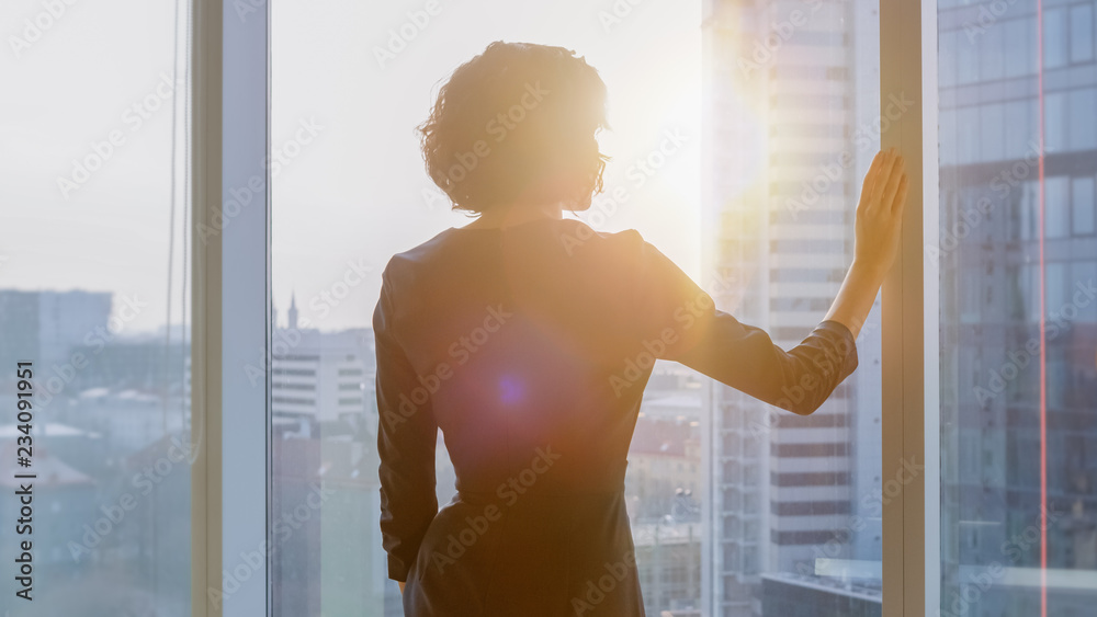 一位成功的女商人穿着引人注目的黑色连衣裙在办公室眺望窗外的照片