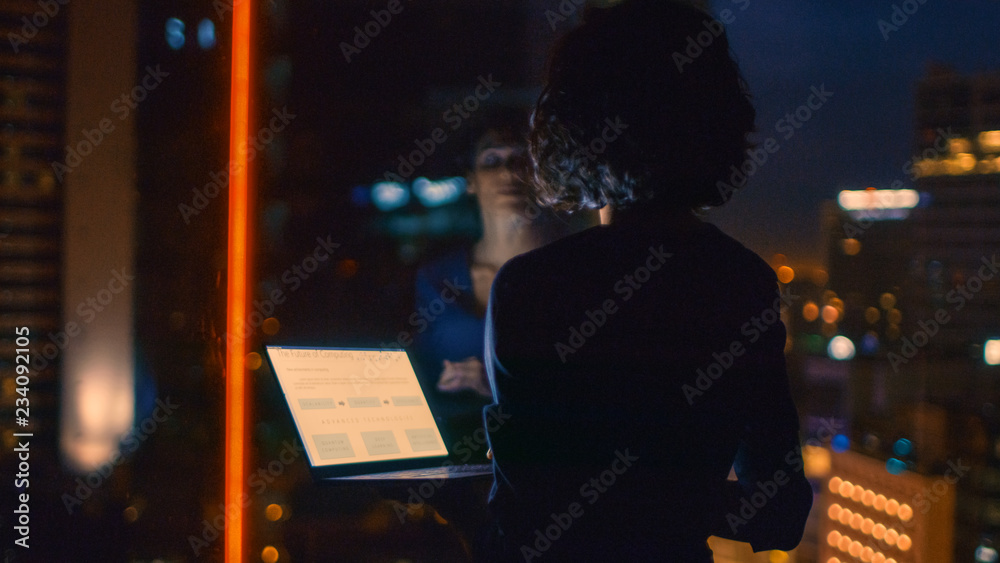 穿着时髦的女商人拿着笔记本电脑，从办公室的窗户向外看。深夜