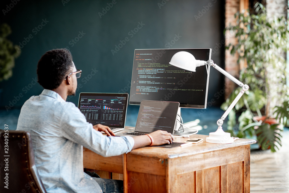 年轻的非洲男性程序员坐在工作场所写程序代码，三个监视器在t中