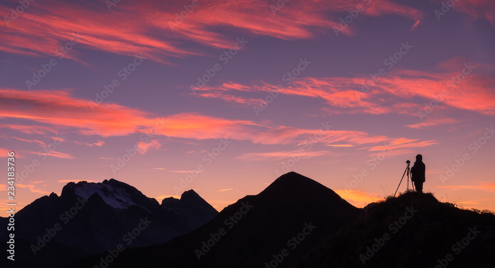 一位摄影师在山峰上的剪影，映衬着岩石和紫色的天空，粉红色的云朵在s处