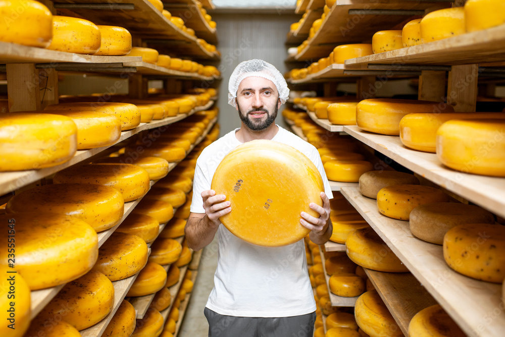 一位英俊的工人拿着奶酪站在装满奶酪轮子的仓库里的肖像