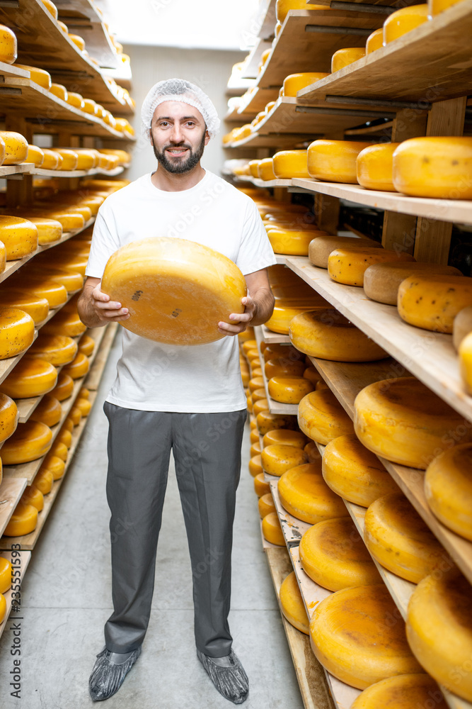 一位英俊的工人拿着奶酪站在装满奶酪轮子的仓库里的肖像