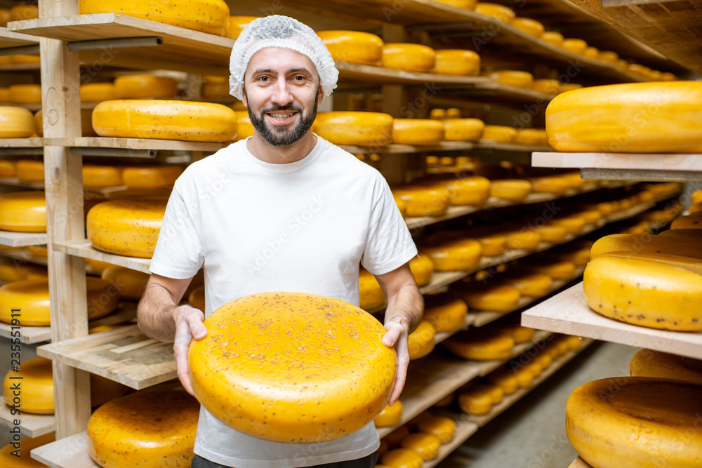 一名工人的画像，他在储藏室里拿着奶酪轮，货架上放满了奶酪。