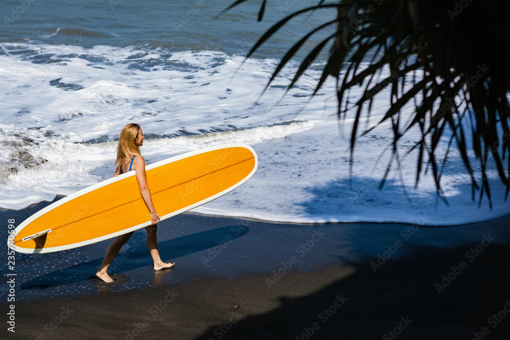 穿着比基尼、带着冲浪板的运动女孩在黑沙滩上行走。年轻的冲浪女子在水池边奔跑
