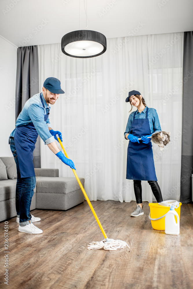Professional cleaners in blue uniform washing floor and wiping dust from the furniture in the living