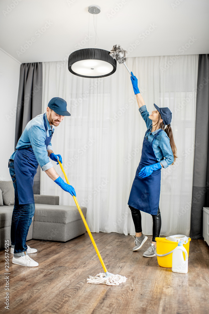穿着蓝色制服的专业清洁工在客厅洗地板、擦灯上的灰尘