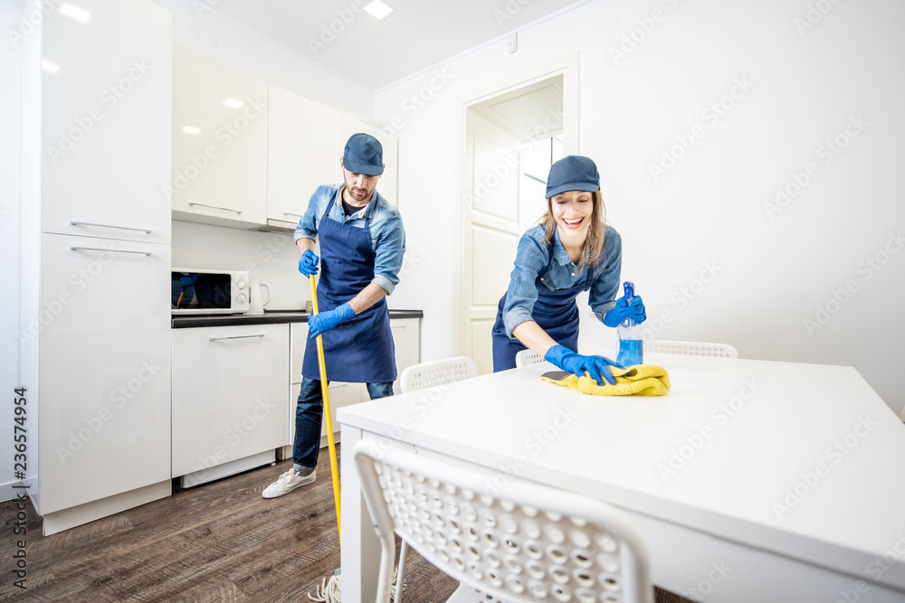 Man and woman as a professional cleaners in uniform washing floor and wiping furniture in the white 