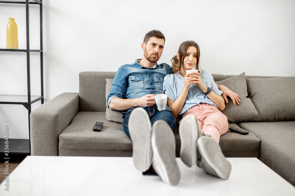 一对年轻夫妇坐在家里的沙发上看无聊的电视，脸上挂着不开心的表情