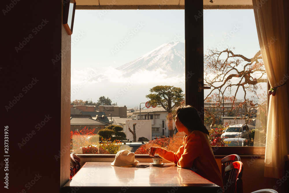 年轻的亚洲女人在酒店的窗户边欣赏富士山的景色，喝咖啡