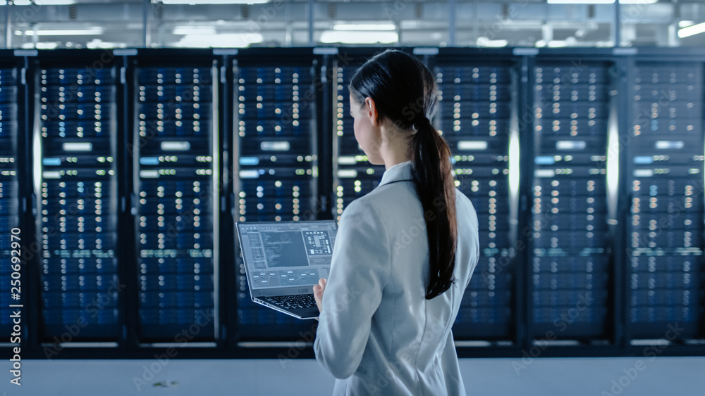 一位女性数据中心IT专家在服务器机架Corrido旁边的笔记本电脑上工作的背影