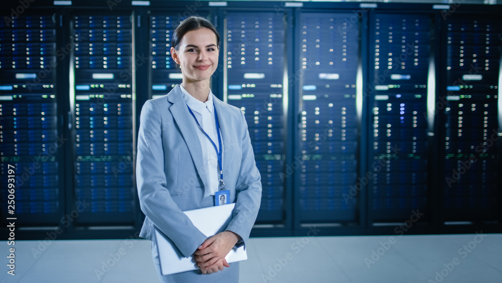 女IT专家站在数据中心服务器机架旁边，拿着笔记本电脑微笑着