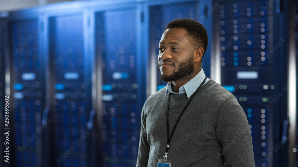 黑人数据中心IT技术员站在服务器机架走廊中间。他在微笑。