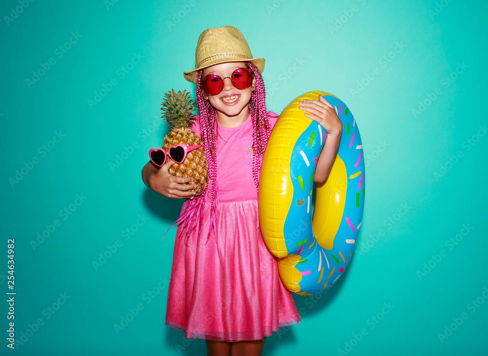 Funny happy child girl in summer pink dress with pineapple and swimming circle on blue background.