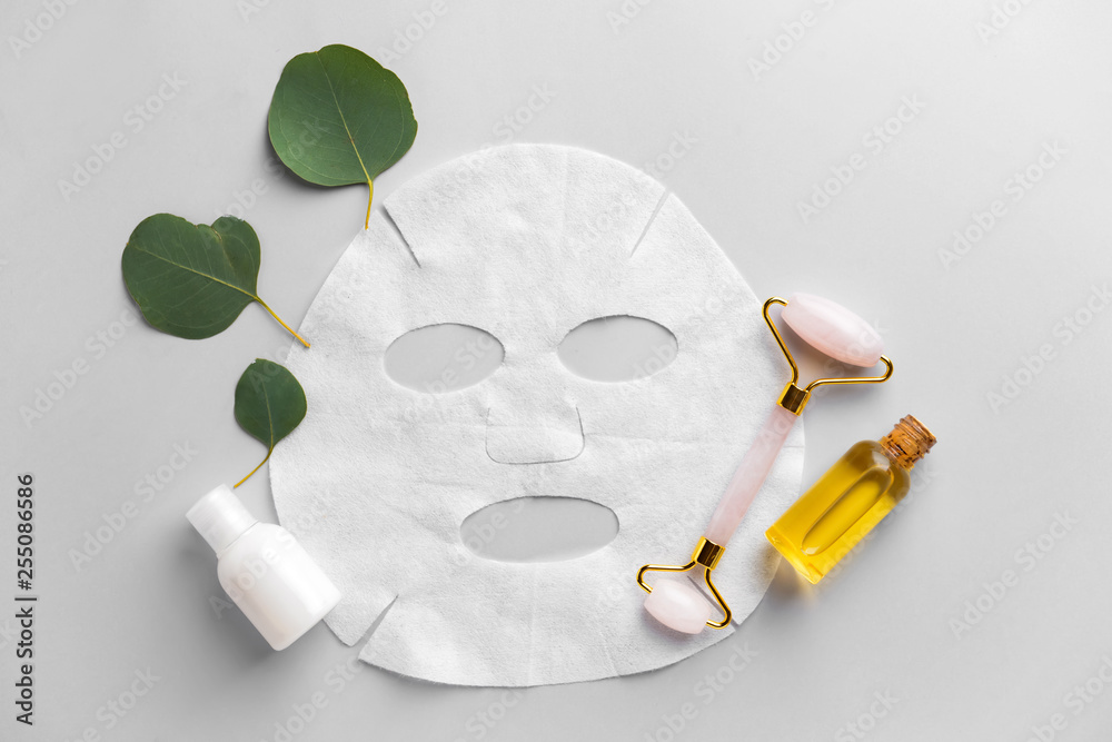 Sheet facial mask with cosmetic products and massage tool on light background