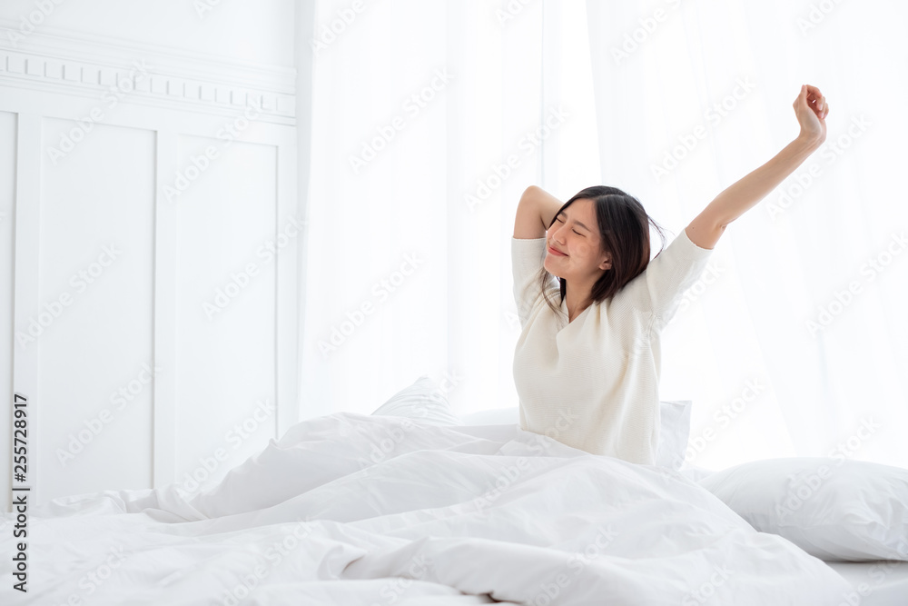 亚洲女人早上醒来，坐在白色的床上伸展身体，感到幸福和新鲜