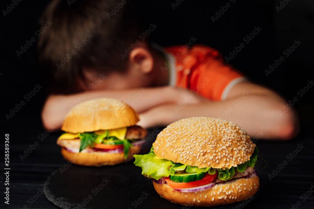悲伤的孩子坐在桌子旁，在美味的汉堡前把头靠在胳膊上。美味的汉堡